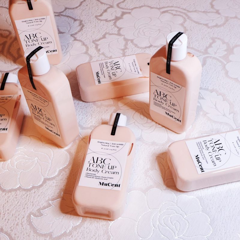 MuCent ABC Tone Up Body Cream products photographed with soft lighting on a floral textured surface